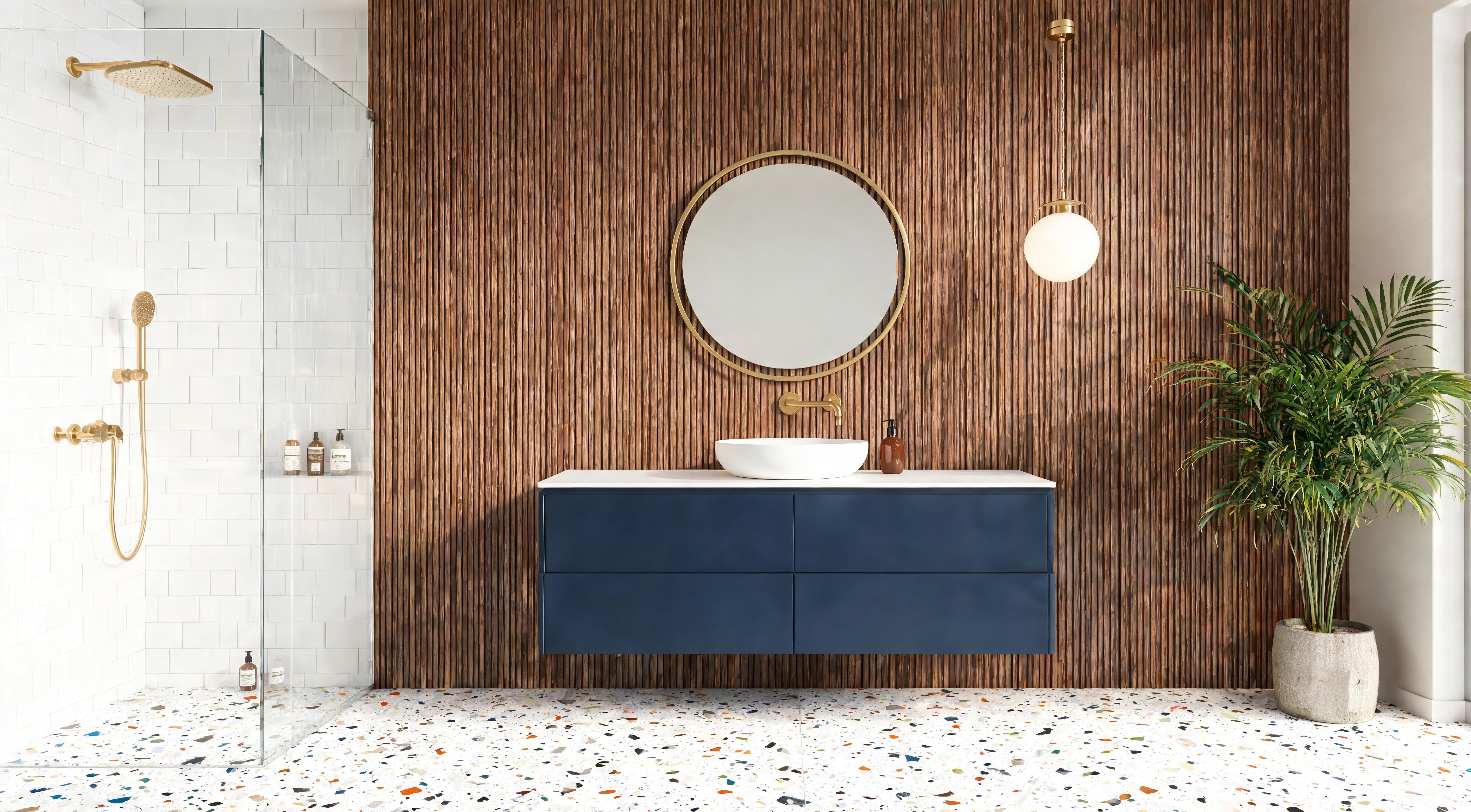 A bathroom with a natural wood fluted panel feature wall, navy vanity unit, round brass mirror and terrazzo floor