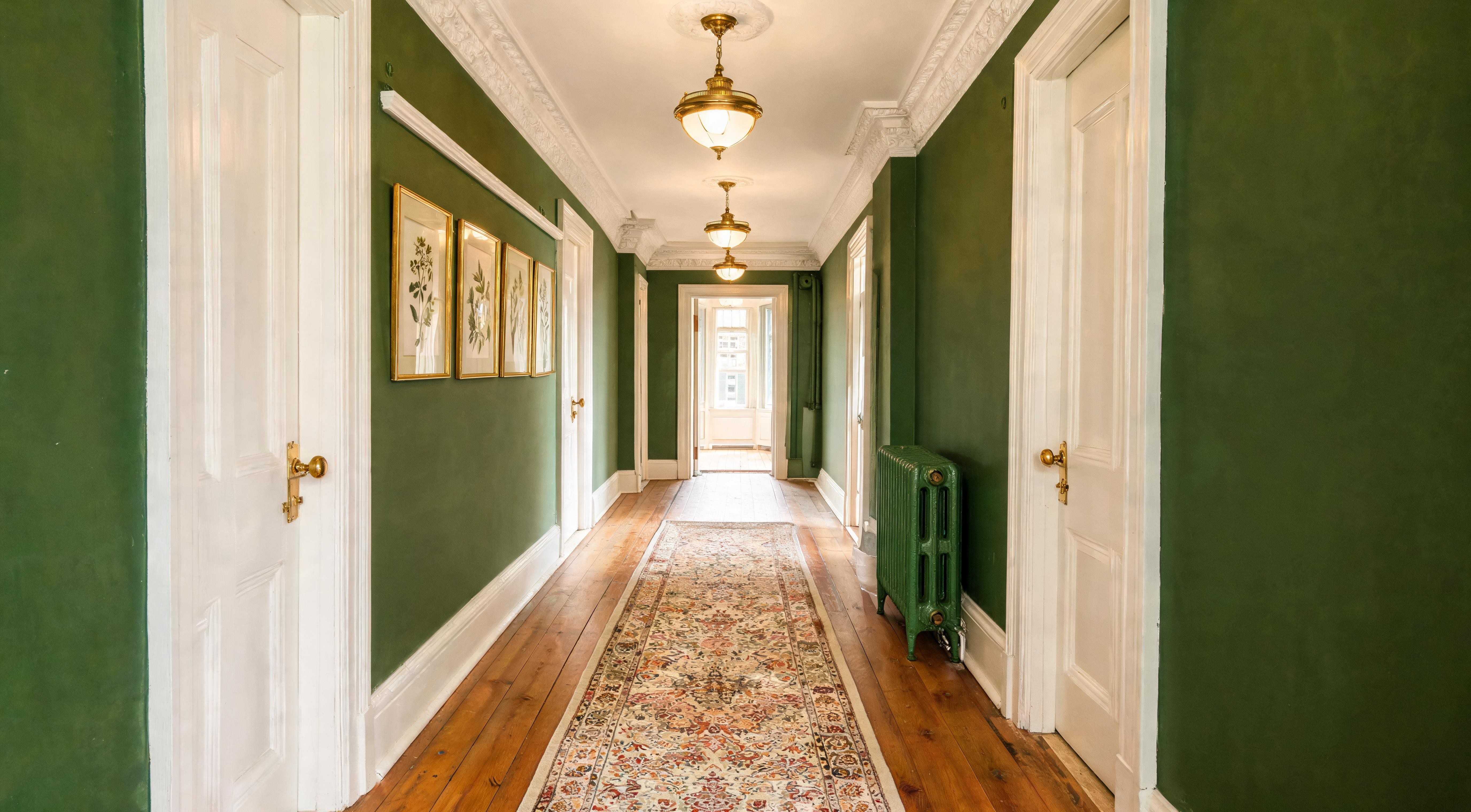 A long narrow hallway painted in deep green with white trim, brass pendant lights, botanical prints and a traditional runner rug